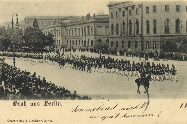 Ein altes Schwarz-Weiß-Foto eines Umzugs in Berlin, Deutschland, mit Menschen auf dem Boden und auf Pferden, einer Statue, Straßenlaternen, Bäumen, Gebäuden mit Fenstern und einem bewölkten Himmel, mit Text am unteren Rand.