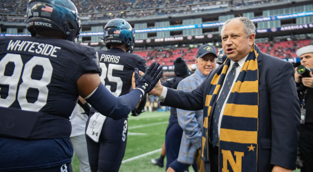 Ein Mann im Anzug und Krawatte gibt einem Fußballspieler die Hand auf einem Feld, umgeben von einer Gruppe von Menschen, mit einer Person, die eine Kamera auf der rechten Seite hält und einem Stadion voller Zuschauer im Hintergrund, das Schilder mit der Aufschrift "Notre Dame Football Coach Mike McCarthy und die Fighting Irish" anzeigt.