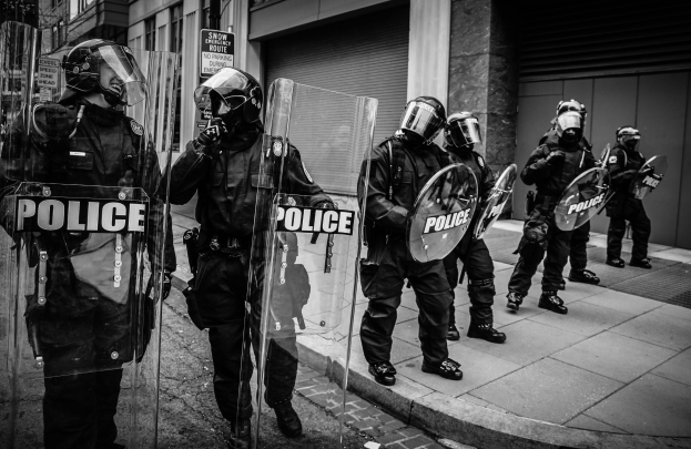 Eine Gruppe von Polizisten in Helmen und mit Schilden mit Text darauf steht auf einem Gehweg vor Gebäuden mit Fenstern, Rollläden und Schildern, alles in Schwarz-Weiß dargestellt.