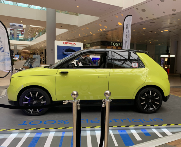 Das erste Wasserstoff-elektrische Auto der Welt, ein gelbes Fahrzeug, steht vor einem Gebäude mit Absperrpollern, Seilen, Bannern, einer Person im Auto, einer Säule, einer Gruppe von stehenden Menschen, Schildern, einem Topf mit einer Pflanze, Deckenleuchten und Lüftern.