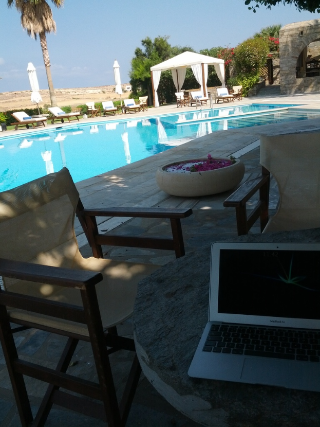 Ein Schwimmbeckenbereich mit Stühlen, einem Tisch mit einem Laptop, einem Topf mit Blumenblüten, Palmen, anderen Bäumen, einem Unterstand und einem Gebäude im Hintergrund unter einem klaren Himmel.