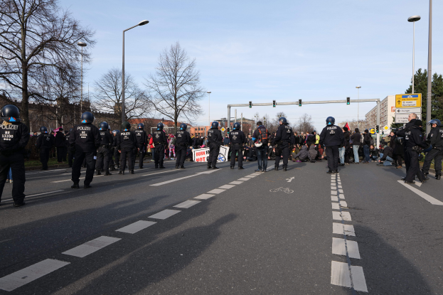 Eine Gruppe von Polizisten in schwarzen Uniformen und Helmen steht an der Seite einer Straße, mit Laternenmöbeln, Ampeln, Bäumen, Gebäuden und einem klaren blauen Himmel im Hintergrund.