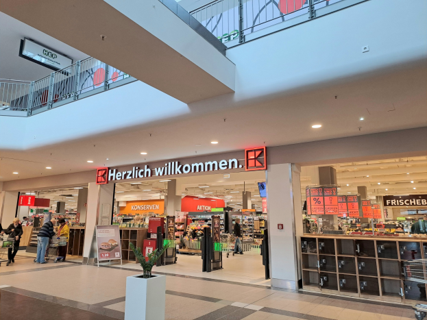 Innenansicht eines Einkaufszentrums mit Menschen, Geschäften, Infotafeln mit der Aufschrift "Herzlich willkommen", Beleuchtung, Geländern und einem Topf mit einer Pflanze unten.
