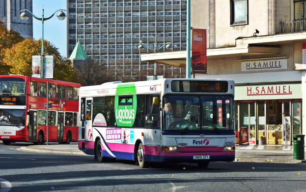 Zwei Busse stehen auf einer Straße mit einem Gebäude, auf dem "H. SAMUEL" steht, und einem weiteren Gebäude dahinter, mit Bäumen und einer Straßenlaterne davor.