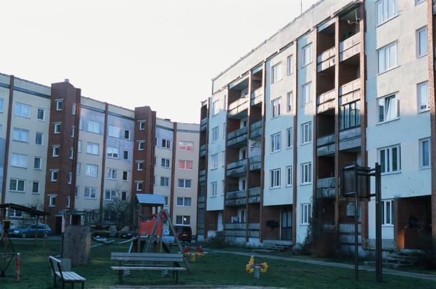 Wohngebäude mit Spielplatz, Gras, Bänken, Schaukeln, Fahrzeugen, Bäumen und einem klaren blauen Himmel im Hintergrund.
