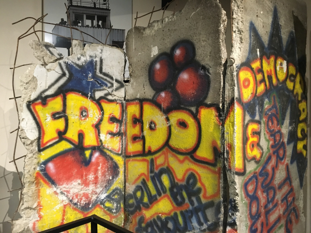 Mauer mit Graffiti, das in fetten schwarzen Buchstaben 'Freiheit und Demokratie' lautet, ein Geländer unten und ein Gebäude mit Fenstern im Hintergrund.