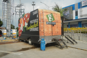 Ein weißer Lastwagen mit einer "Teilen Sie eine positiv überladene Erfahrung"-Aufschrift parkt vor einem Gebäude mit Stufen, einem Zylinder, Geländern, Strommasten, Laternen, Bäumen und einem bewölkten Himmel im Hintergrund.