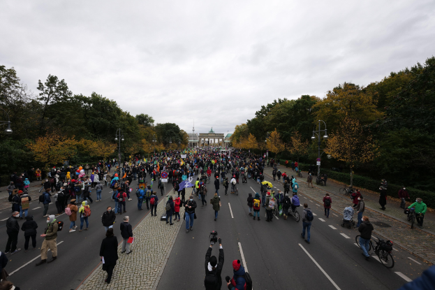 Große Gruppe von Menschen, die eine Straße mit Bäumen und Laternen entlanggehen, während sie an einer Protestdemo teilnehmen, mit einem Gebäude und einem klaren Himmel im Hintergrund.