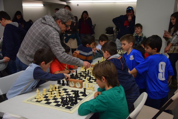 Kinder spielen Schach in einem Wettbewerb, während Erwachsene von hinten und seitlich zuschauen.