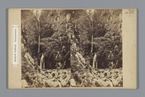 Schwarzes und weißes Foto eines Wasserfalls, der eine felsige Klippe herunterfließt und von Bäumen umgeben ist, mit einem Stempel auf der linken Seite.