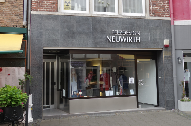Ein Schaufenster mit Glaswänden und -türen, das Puppen in Kleidern zeigt, Topfpflanzen und ein Schild mit der Aufschrift "Pelzdesign Neuwirth in Amsterdam".