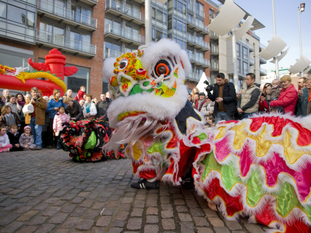 Ein farbenfrohes chinesisches Neujahrsfest in Amsterdam mit einer Löwentanzvorstellung vor einer Zuschauermenge, einige halten Kameras, vor dem Hintergrund von Gebäuden, Laternenmasten und einem klaren blauen Himmel.