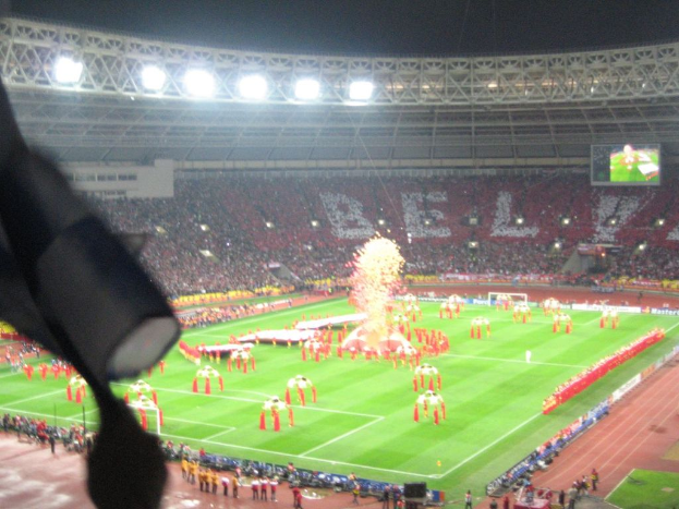 Ein Stadion mit Flutlicht, Eisengittern, Zuschauern, stehenden Personen, Werbetafeln, Bildschirmen, einer Überwachungskamera und verschiedenen Dekorationen einschließlich Flaggen.