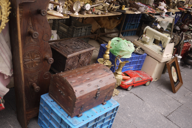 Eine Flohmarktszene mit Holzboxen, einem Spielzeugauto, einer Nähmaschine, einem Foto Rahmen, einer Uhr und anderen Gegenständen auf Tischen.