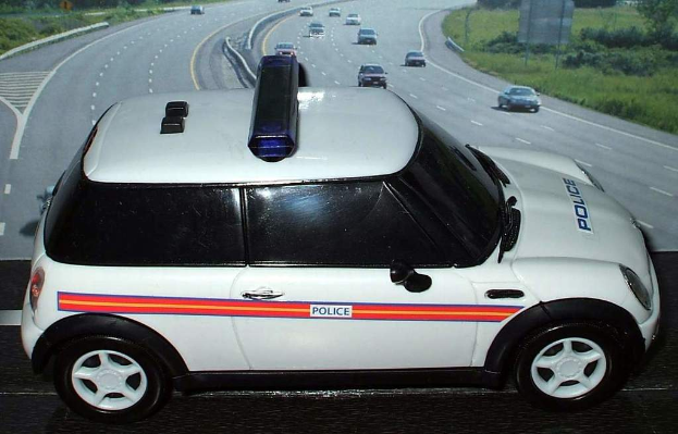 Ein weißes Polizeifahrzeug steht im Vordergrund mit mehreren Fahrzeugen, die sich auf der Straße dahinter bewegen, umgeben von Gras und Bäumen auf beiden Seiten.