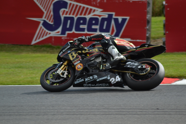 Eine Person in Helm und Schuhen fährt ein Motorrad auf einer Rennstrecke, die von Gras umgeben ist, mit einer Tafel mit Text im Hintergrund.