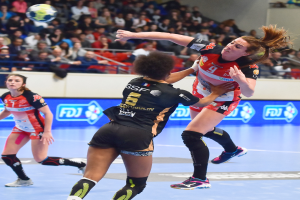 Eine Gruppe von Frauen spielt Handball auf einem Feld, während Zuschauer von Stühlen, Geländern und einer Wand im Hintergrund zusehen.