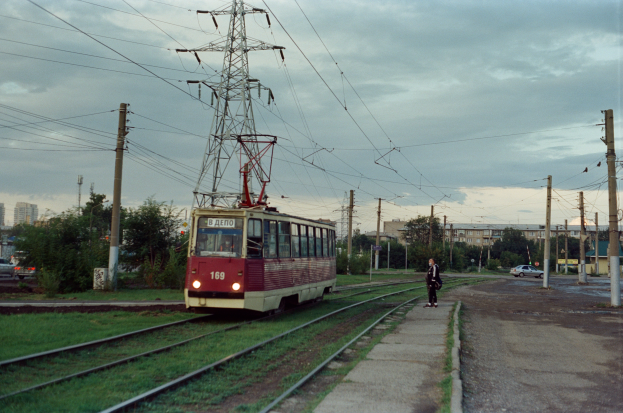 Eine rote und weiße Straßenbahn fährt auf Schienen neben einer Stromleitung, mit einer Person auf dem Bahnsteig; im Hintergrund sind Strommasten, Fahrzeuge, Bäume, Gebäude und ein bewölkter Himmel zu sehen.